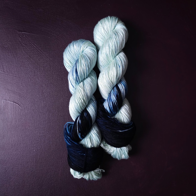 May include: Two skeins of yarn in shades of light blue, navy, and black. The yarn is twisted into a rope-like shape, with the colors transitioning from light to dark. The background is a dark purple.