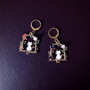 May include: A pair of gold-tone earrings featuring a white cat silhouette design. The cat is seated within a moon and star archway adorned with green leaves and a pearl accent.
