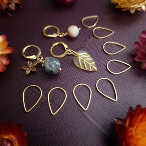 May include: A set of gold-toned stitch markers with various charms, including a leaf, a flower, and a green agate stone. The markers are shaped like teardrops and are arranged on a purple surface.