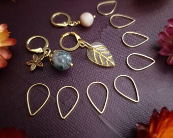 May include: A set of gold-toned stitch markers with various charms, including a leaf, a flower, and a green agate stone. The markers are shaped like teardrops and are arranged on a purple surface.