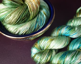 Könnte beinhalten: Eine Garnrolle in Grün, Blau und Weiß mit goldenen Sprenkeln. Das Garn ist um sich selbst gewickelt und in einer blauen und goldenen Schale platziert. Das Garn ist auch in einer losen Spirale zu sehen.
