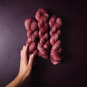 May include: Three skeins of hand-dyed yarn in shades of red and pink, with subtle variations in color. The yarn is arranged vertically against a dark purple background. The image is well-lit, showcasing the texture and color variations of the yarn.