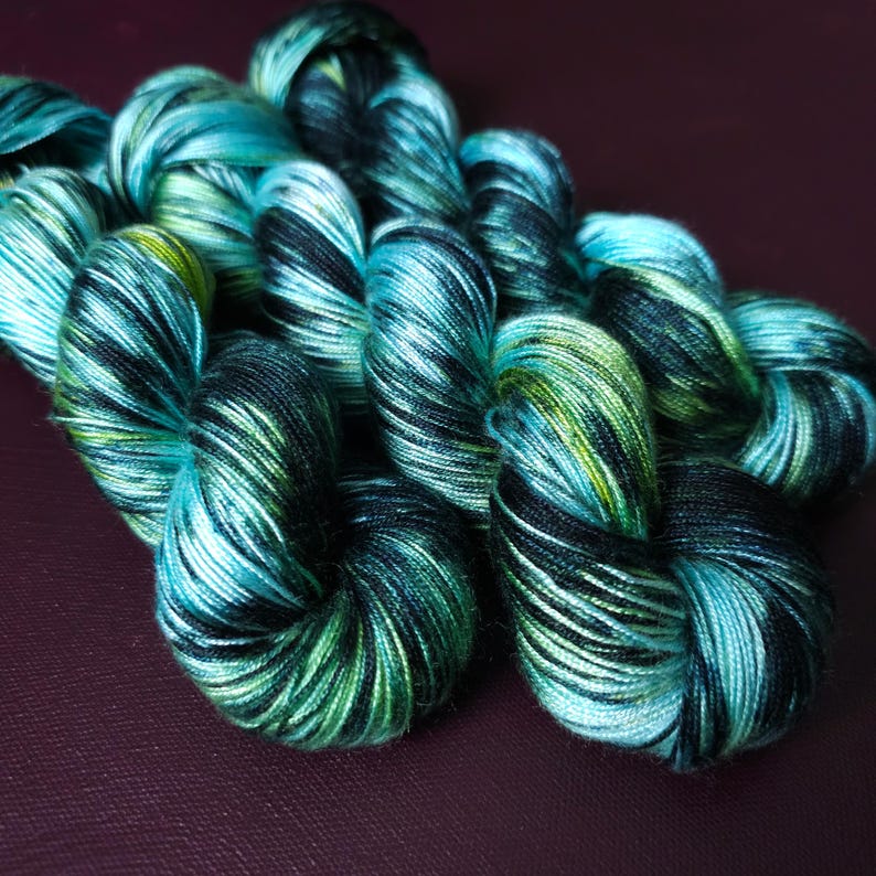 May include: Close-up of several skeins of hand-dyed yarn in shades of teal, green, and black. The yarn is tightly wound, showing a variegated color pattern. The yarn is likely for knitting or crochet projects.