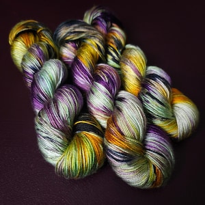 Könnte beinhalten: Drei Knäuel handgefärbtes Garn in den Farben Grün, Lila, Schwarz und Gold. Das Garn liegt in einem lockeren Haufen.