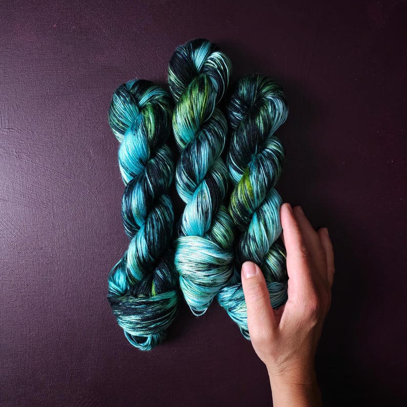 May include: Three skeins of hand-dyed yarn in shades of teal, green, and black. The yarn is arranged on a dark purple surface. The yarn is a soft, textured material, perfect for knitting or crocheting projects.