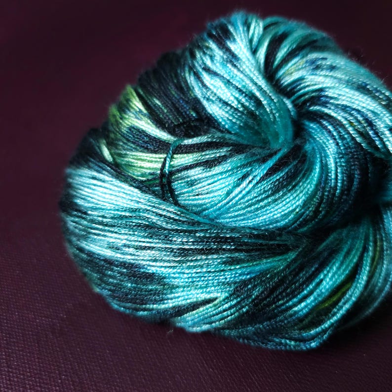 May include: A close-up shot of a skein of yarn. The yarn is a blend of teal, turquoise, black, and green. The yarn is tightly wound and has a slight sheen. The background is a solid, dark purple color.