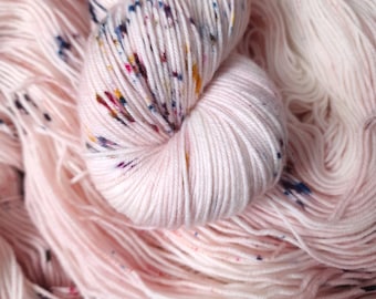 May include: A skein of soft, white yarn with colorful flecks throughout. The yarn is loosely wound and has a delicate, airy texture.