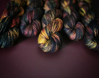 May include: Close-up of several skeins of yarn in a dark blue, black, yellow, and orange colour combination. The yarn is arranged in a semi-circle on a dark purple surface.