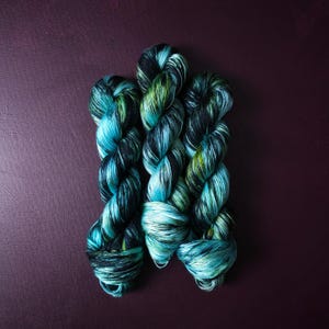 May include: Three skeins of hand-dyed yarn in shades of teal, green, and black. The yarn is twisted into a rope-like structure, with the colors blending together. The background is a dark purple.