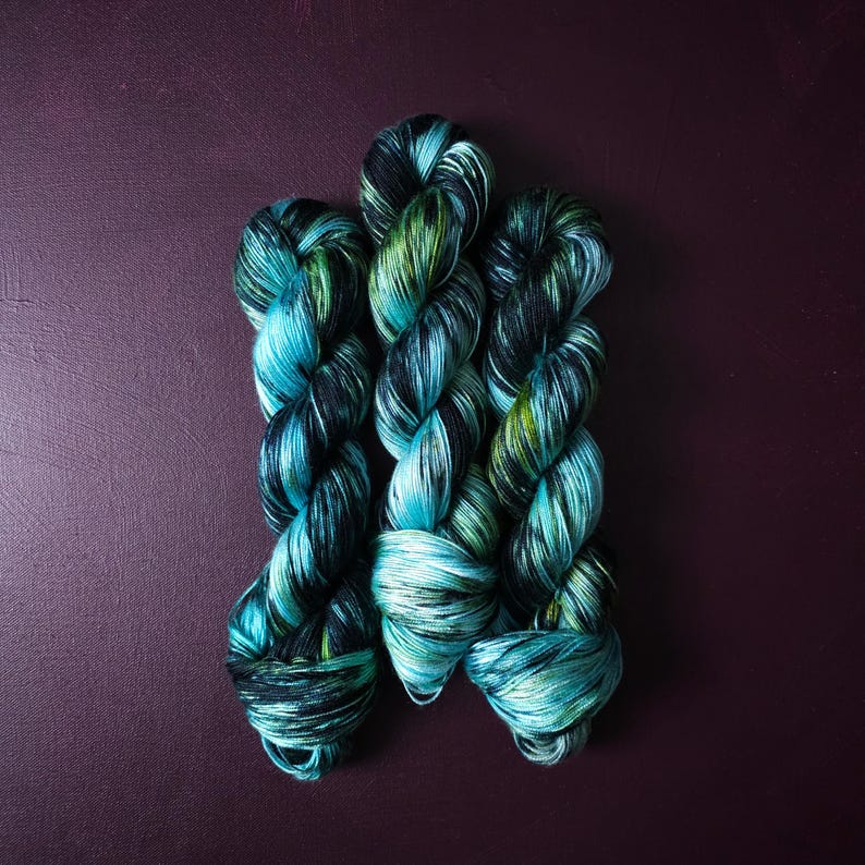 May include: Three skeins of hand-dyed yarn in shades of teal, green, and black. The yarn is twisted into a rope-like structure, with the colors blending together. The background is a dark purple.