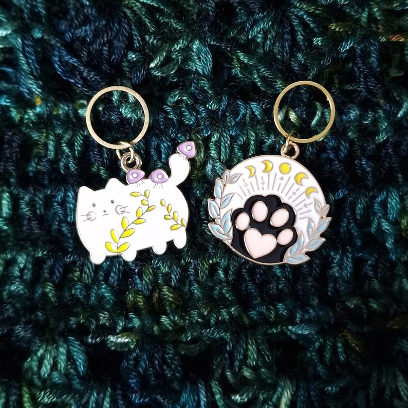 May include: Two enamel stitch markers with gold-coloured rings. One is a white cat with purple mushrooms and yellow leaves. The other is a white circle with a black paw print, a pink heart, and a crescent moon design.
