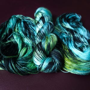May include: Close-up of three skeins of hand-dyed yarn in shades of teal, green, and black. The yarn is arranged in soft, flowing bundles, with visible strands and subtle color variations. The background is a deep, muted purple.