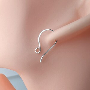 May include: A silver-colored earring with a curved design. The earring is a simple, minimalist style, featuring a hook that curves into a small loop. The earring is shown on a light skin tone.