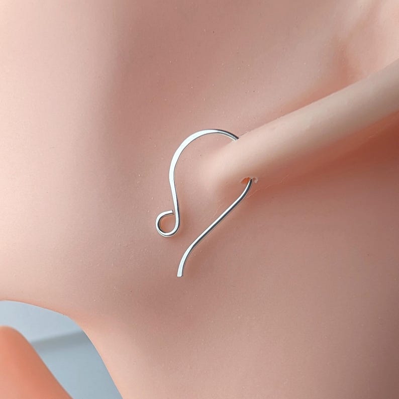 May include: A silver-colored earring with a curved design. The earring is a simple, minimalist style, featuring a hook that curves into a small loop. The earring is shown on a light skin tone.
