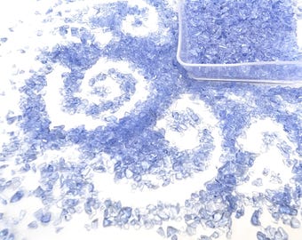 May include: A close-up shot of light blue crushed glass pieces scattered on a white surface, forming a spiral design. A clear plastic container holds more of the glass. The glass pieces are small and irregular in shape.