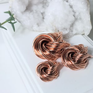 May include: Three copper-colored wire knot sculptures of varying sizes are displayed on a white surface. The sculptures have a twisted, interwoven design. A white crystal geode and a sprig of greenery are in the background.