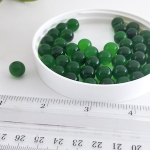 May include: A collection of small, translucent green glass marbles inside a white container. A single marble rests outside the container. A clear ruler with inch markings is in the foreground.