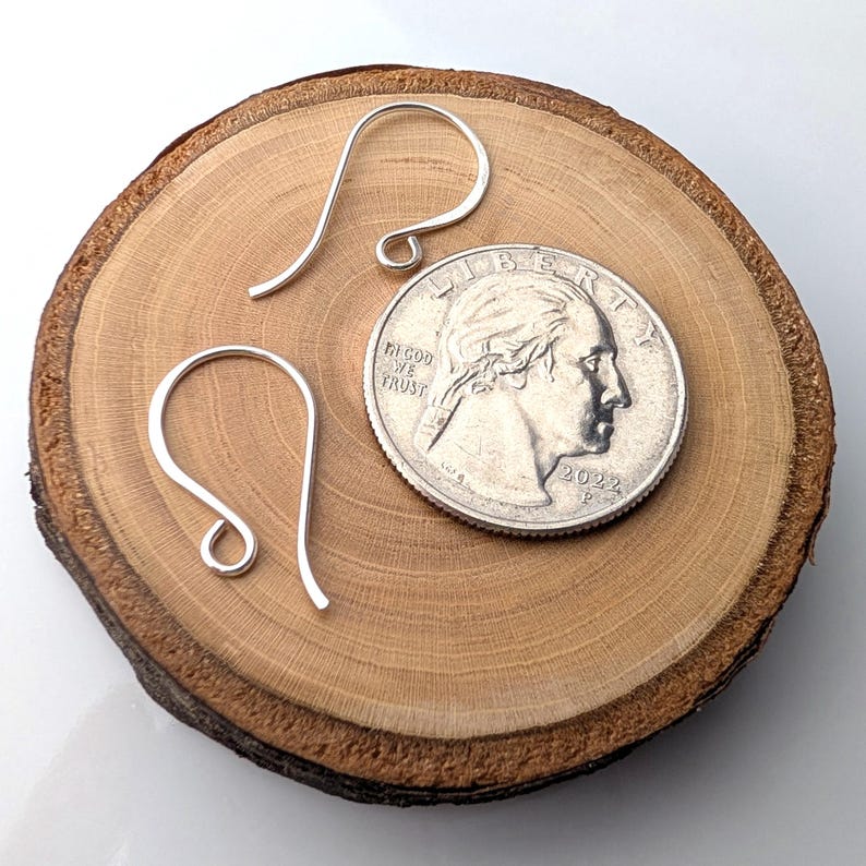 May include: Two silver-colored earring hooks are displayed on a wooden surface next to a U.S. quarter. The hooks are curved and have a simple, elegant design. The quarter is dated 2022 and provides a size reference.