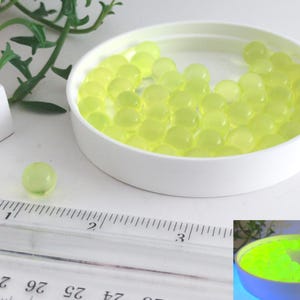 May include: A collection of translucent, light green gel balls inside a white container. One gel ball sits on a white surface next to a ruler. The gel balls have a glowing appearance. The image also includes a small plant.