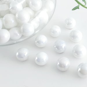 May include: A collection of white, iridescent glass marbles, some spilling from a clear glass container. The marbles are scattered on a white surface, with a few green leaves in the background. These decorative marbles are ideal for home decor.