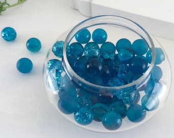 May include: A clear glass bowl filled with various sizes of blue glass marbles. Some marbles are solid, while others have a cracked, textured appearance. Several marbles are scattered around the bowl on a white surface.