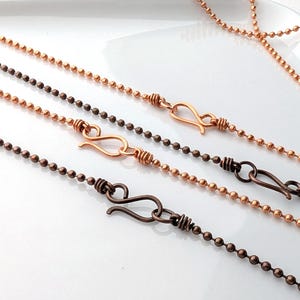 May include: Close-up of four ball chain necklaces in copper and bronze tones. Each necklace features a unique hook clasp. The chains are displayed on a white surface, showcasing the different metallic finishes and textures.