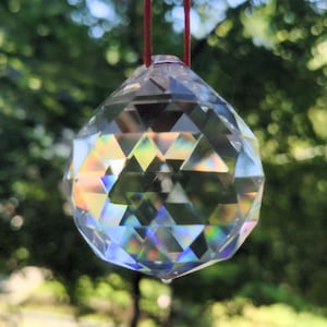 May include: A clear, faceted crystal ball hanging from a red string. The crystal reflects a rainbow of colors, including yellow, green, and blue, against a blurred green background of foliage.