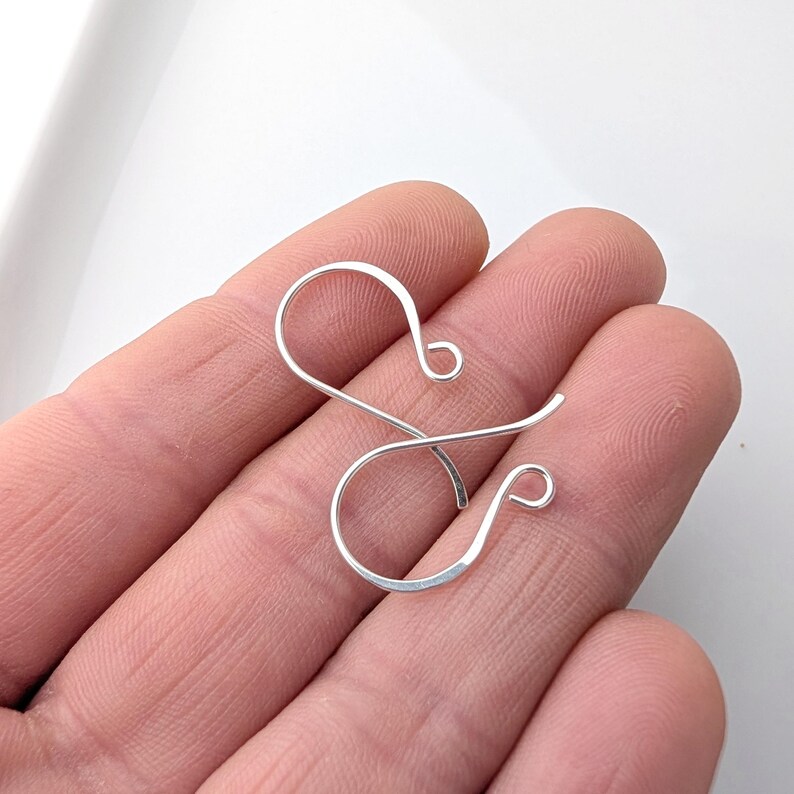 May include: A single, silver-colored earring hook, crafted from thin wire, is held in a hand. The hook features a looped design at both ends, with a curved, open shape for insertion into the ear. The earring hook is shown against a neutral background.