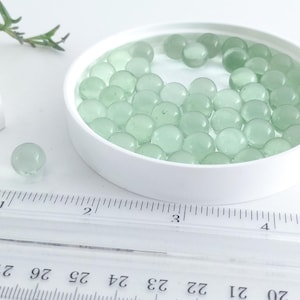 May include: A collection of translucent, pale green glass spheres inside a white, round container. One sphere sits separately. A ruler with inch markings is in the foreground. The spheres are approximately 1 inch in diameter.