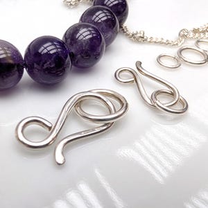 May include: A collection of jewelry components including several deep purple, spherical amethyst beads strung together, accompanied by two silver-toned metal S-hook clasps and several small silver-toned jump rings arranged on a white surface.
