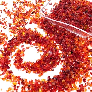 May include: A close-up shot of scattered, crushed, red and orange glass pieces. A clear, rectangular scoop is filled with the glass pieces. The glass pieces are scattered on a white surface.