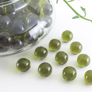 May include: A clear glass bowl filled with dark green glass marbles. Several green marbles are scattered on a white surface. A green plant stem is in the background. Decorative glass marbles.