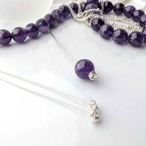 May include: A set of silver hair pins featuring a single round purple amethyst bead on one pin and coiled silver wire ends on the others. A strand of round purple amethyst beads and silver chain is positioned in the background of the hair pins.