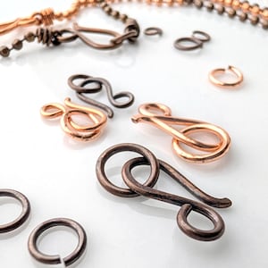 May include: A selection of copper and bronze jewellery findings, including clasps, jump rings, and chain. The clasps have a curved design, and the jump rings are circular. The chain is made of small, round beads. The findings are scattered on a white surface.
