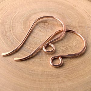 May include: Two copper-colored earring hooks are displayed against a wooden background. The hooks have a curved design with a looped bottom and a pointed top. The metal has a warm, reddish-brown hue.