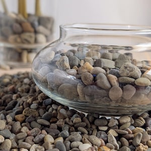 May include: A clear glass bowl filled with smooth, gray and brown river rocks. The bowl sits atop a bed of similar stones, with another glass container holding similar rocks in the background. The scene evokes a natural, earthy aesthetic.