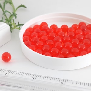 May include: A white, round container filled with numerous small, translucent red spheres. One sphere sits separately. A ruler with inch markings is in the foreground. The objects are on a white surface.
