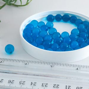May include: A white container filled with numerous translucent blue gel beads. One bead sits separately, next to the container. A clear ruler with inch markings is in the foreground. The beads are a vibrant, eye-catching blue.