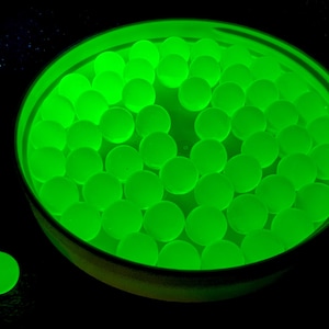 May include: A container filled with numerous glowing green spheres, with one sphere separate from the container. The spheres and container have a bright, neon green color, set against a black background.