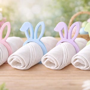 Puede incluir: Cuatro servilleteros pastel en forma de orejas de conejo de Pascua. Los servilleteros son rosas, azules, morados y verdes. Cada anillo rodea una servilleta blanca enrollada. Una cesta de huevos de Pascua está en el fondo.