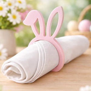 Puede incluir: Un servilletero rosa con forma de orejas de conejo rodea una servilleta blanca enrollada. El anillo tem&aacute;tico de Pascua est&aacute; hecho de un material rosa claro. El fondo presenta una escena borrosa al aire libre con flores y una cesta.