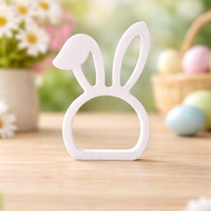 Puede incluir: Un objeto decorativo blanco en forma de orejas de conejo, posiblemente un servilletero o decoraci&oacute;n de Pascua, se encuentra sobre una superficie de madera. El fondo incluye flores borrosas, huevos de Pascua y una cesta, sugiriendo un tema primaveral o de Pascua.