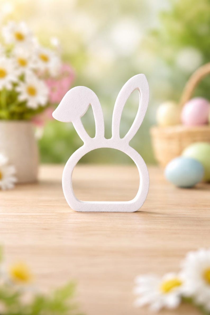 Puede incluir: Un objeto decorativo blanco en forma de orejas de conejo, posiblemente un servilletero o decoraci&oacute;n de Pascua, se encuentra sobre una superficie de madera. El fondo incluye flores borrosas, huevos de Pascua y una cesta, sugiriendo un tema primaveral o de Pascua.