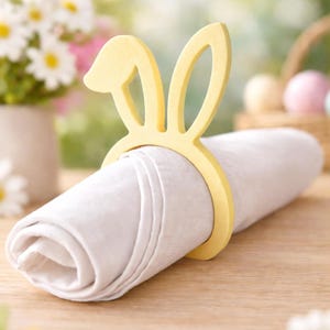 Puede incluir: Una servilleta de lino blanco enrollada sujeta por un servilletero amarillo con forma de orejas de conejo. El servilletero es de madera y tiene un dise&ntilde;o recortado. El fondo est&aacute; borroso, con flores y huevos de Pascua visibles.