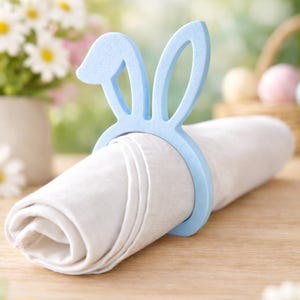 Puede incluir: Un servilletero azul claro con forma de orejas de conejo rodea una servilleta blanca enrollada. El anillo de Pascua est&aacute; hecho de un material s&oacute;lido y se encuentra sobre una superficie de madera. Hay flores en el fondo.