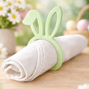 Puede incluir: Un servilletero verde claro con forma de orejas de conejo rodea una servilleta de lino blanco enrollada. El anillo tem&aacute;tico de Pascua est&aacute; hecho de un material s&oacute;lido y se asienta sobre una superficie de madera. Fondo suave con flores y huevos de Pascua.