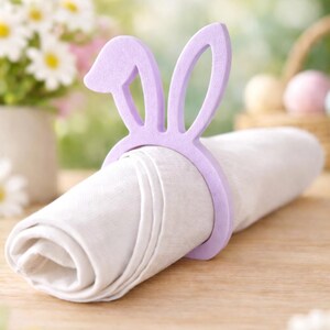Puede incluir: Una servilleta de lino blanco enrollada sujeta por un servilletero de orejas de conejo lila. La decoraci&oacute;n de mesa con tem&aacute;tica de Pascua est&aacute; sobre una superficie de madera, con flores y una cesta.