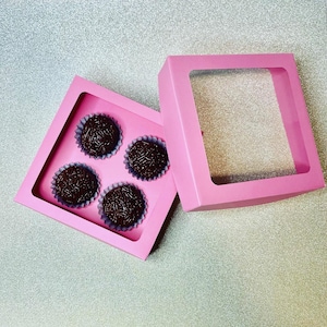 Könnte beinhalten: Vier Schokoladentrüffel mit Streuseln in einer rosa Box mit einem durchsichtigen Fenster.
