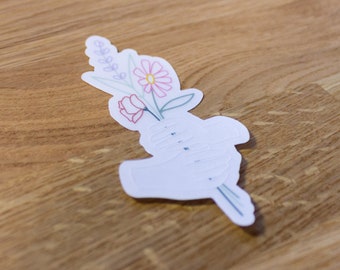 May include: A white sticker with a hand holding a bouquet of flowers. The flowers are pink, red, and purple. The sticker is on a wooden surface.