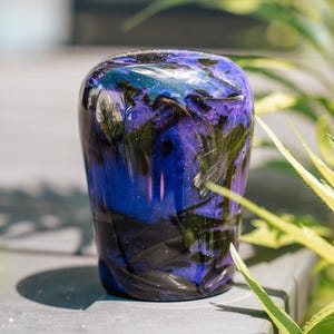 Puede incluir: Objeto decorativo de vidrio, hecho a mano, con una parte superior redondeada y una base cilíndrica. El vidrio presenta una mezcla de azules profundos, púrpuras y negro, con un efecto moteado similar a una galaxia. El objeto mide unos 10 cm de alto.